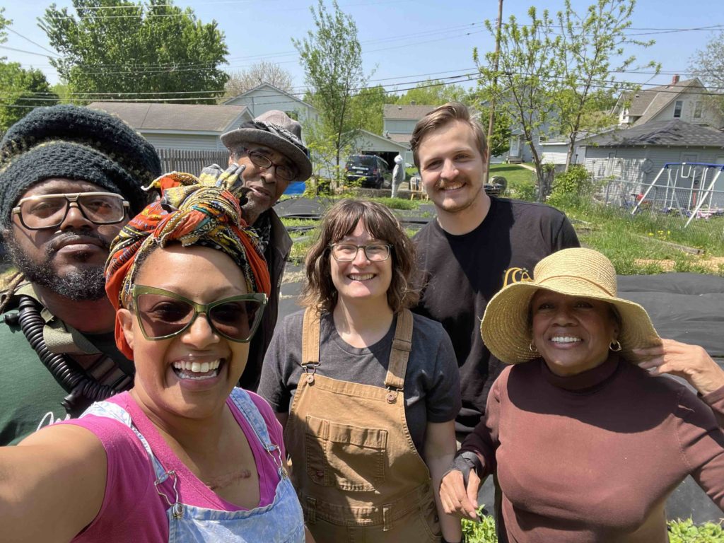 Six people smile for a selfie, outdoors at R. Roots Garden. They are wearing overalls, hats, and other gardening attire.