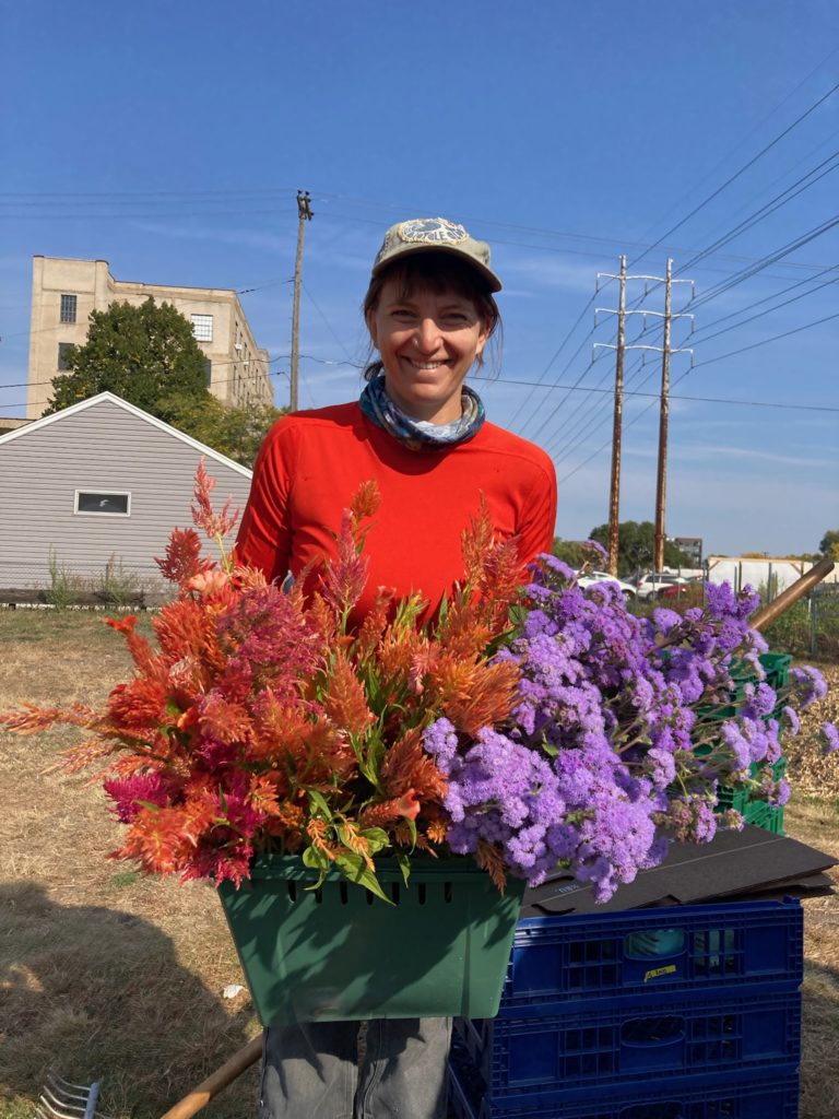 A woman with a hat and bandana stands smiling, holding flowers she grew on her urban farm. There are power lines and buildings in the background.